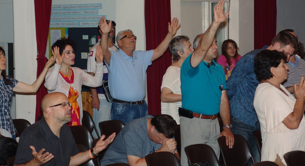 An Albanian man lifts both arms in worship beneath a Matthew 28 banner as the congregation prays