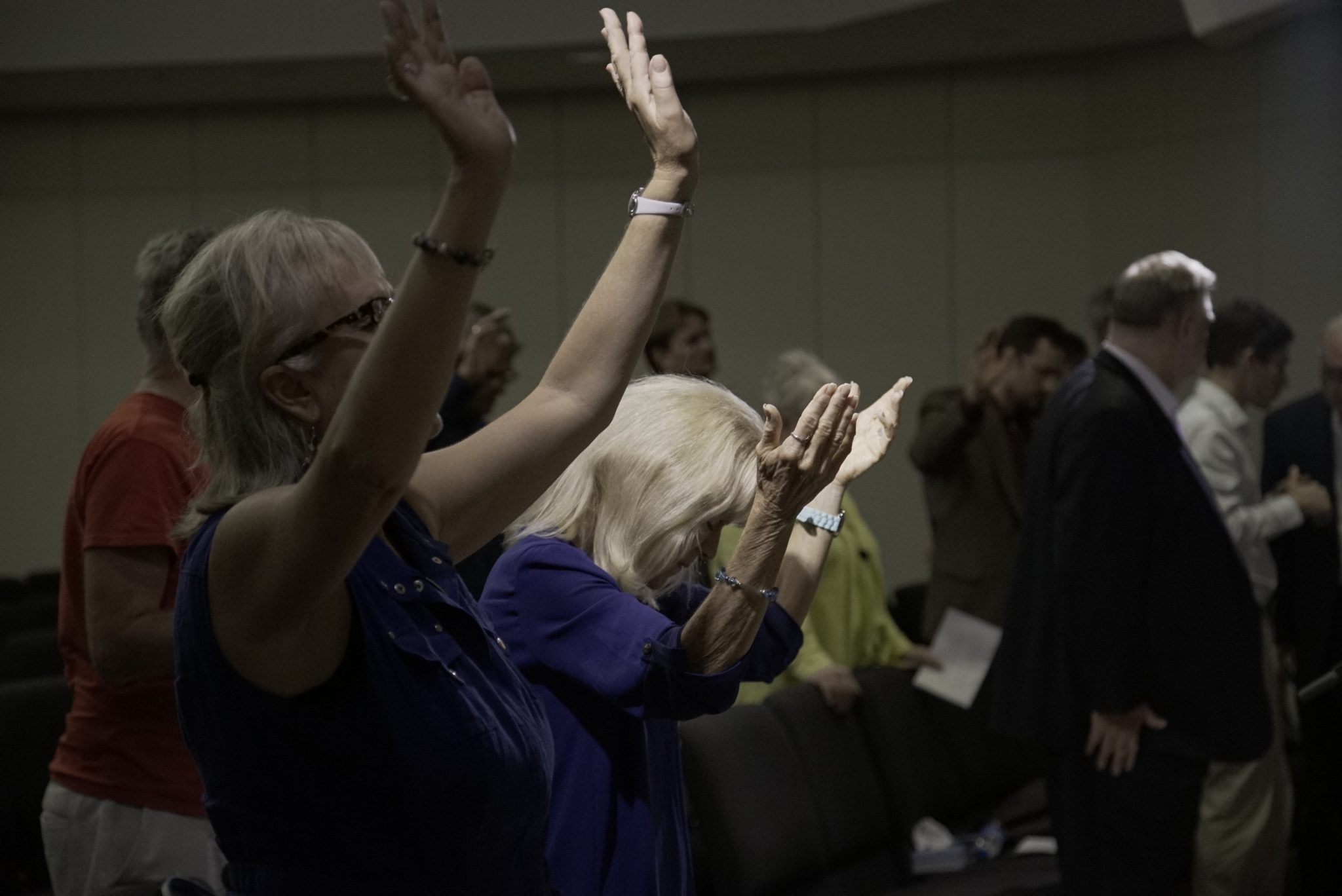 Two women lift open hands in worship as a congregation stands behind them at a closing service.