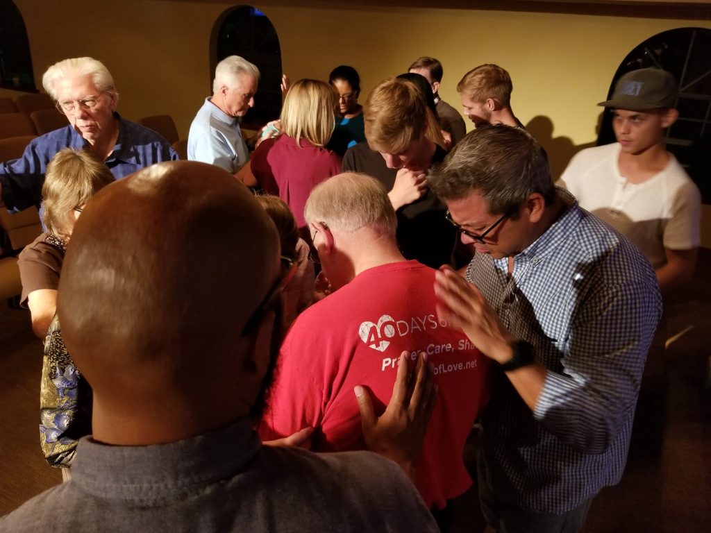People stand in a close circle with hands on one another's shoulders, praying together at a 40 Days of Love gathering.