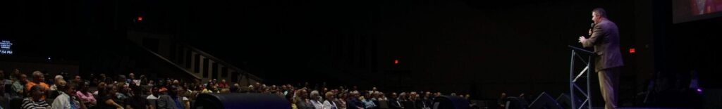 A speaker addresses a seated congregation from a lectern across a wide, purple-lit stage.