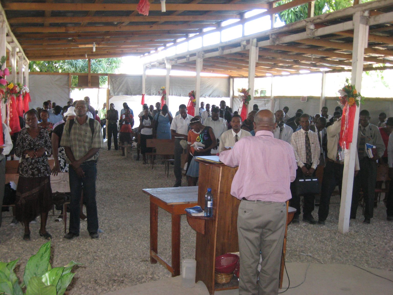 An open-air church congregation standing together in worship under a wooden shelter.