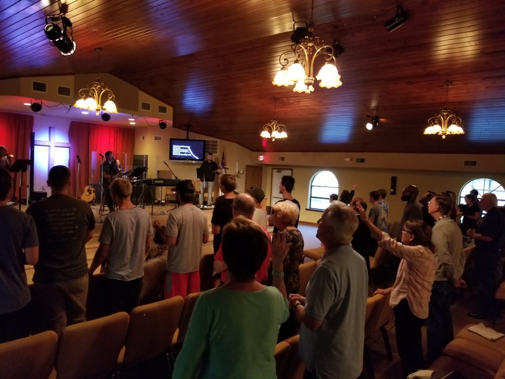 A congregation stands together in worship in a church sanctuary, some with hands raised.