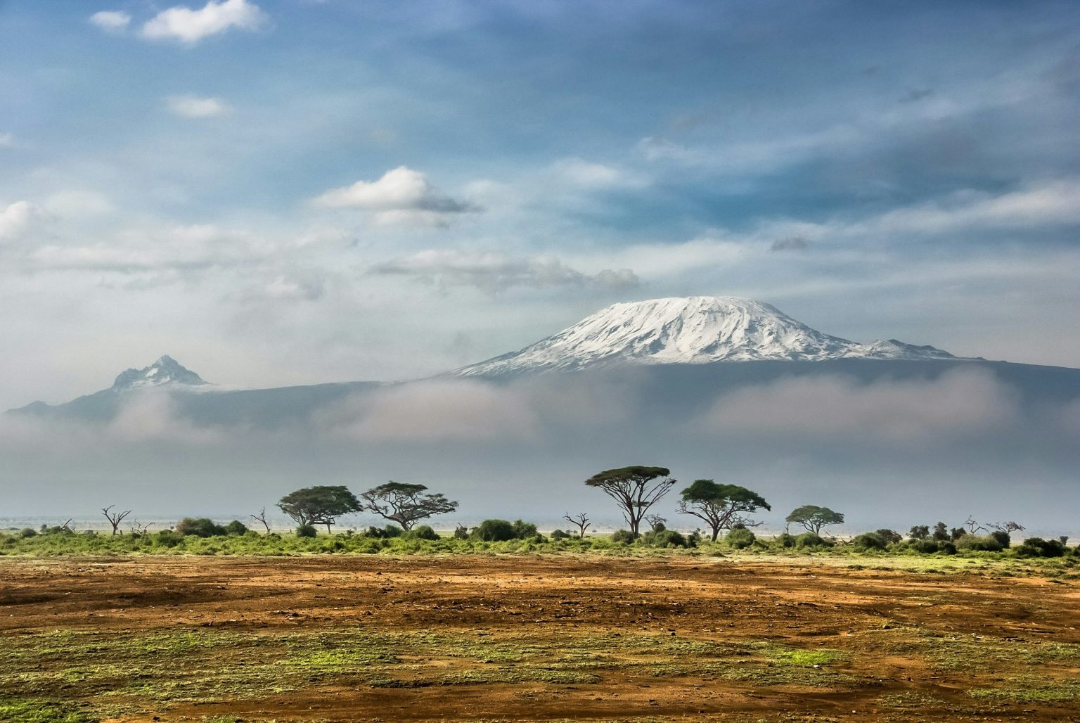 Snow-capped Mount Kilimanjaro rising above an acacia-dotted savanna plain under a wide, cloud-banded sky.