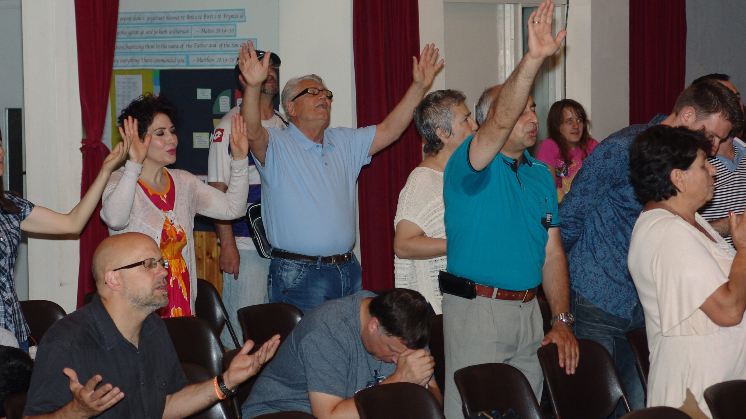 An Albanian man lifts both arms in worship beneath a Matthew 28 banner as the congregation prays