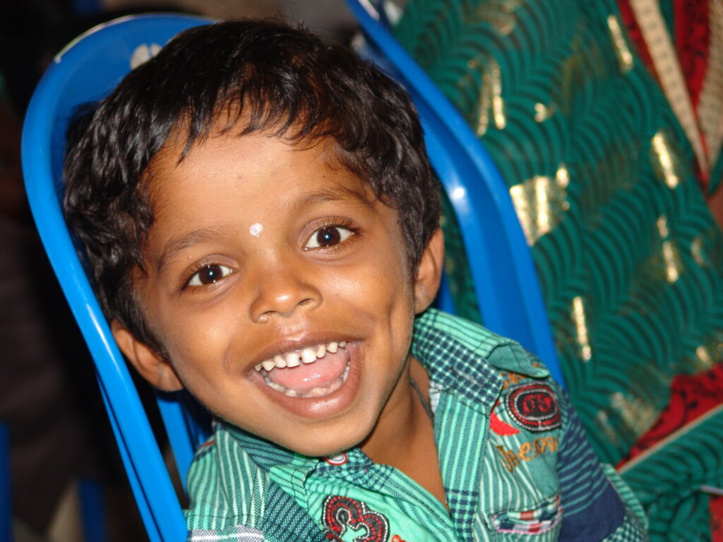 A young boy in a green plaid shirt laughs from a blue plastic chair at an outreach event in India.