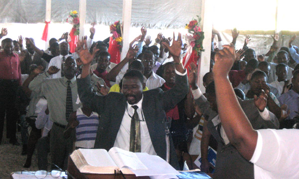 A preacher stands at a lectern with an open Bible, one arm raised, leading a packed congregation under a white tent with dozens of hands lifted in worship.