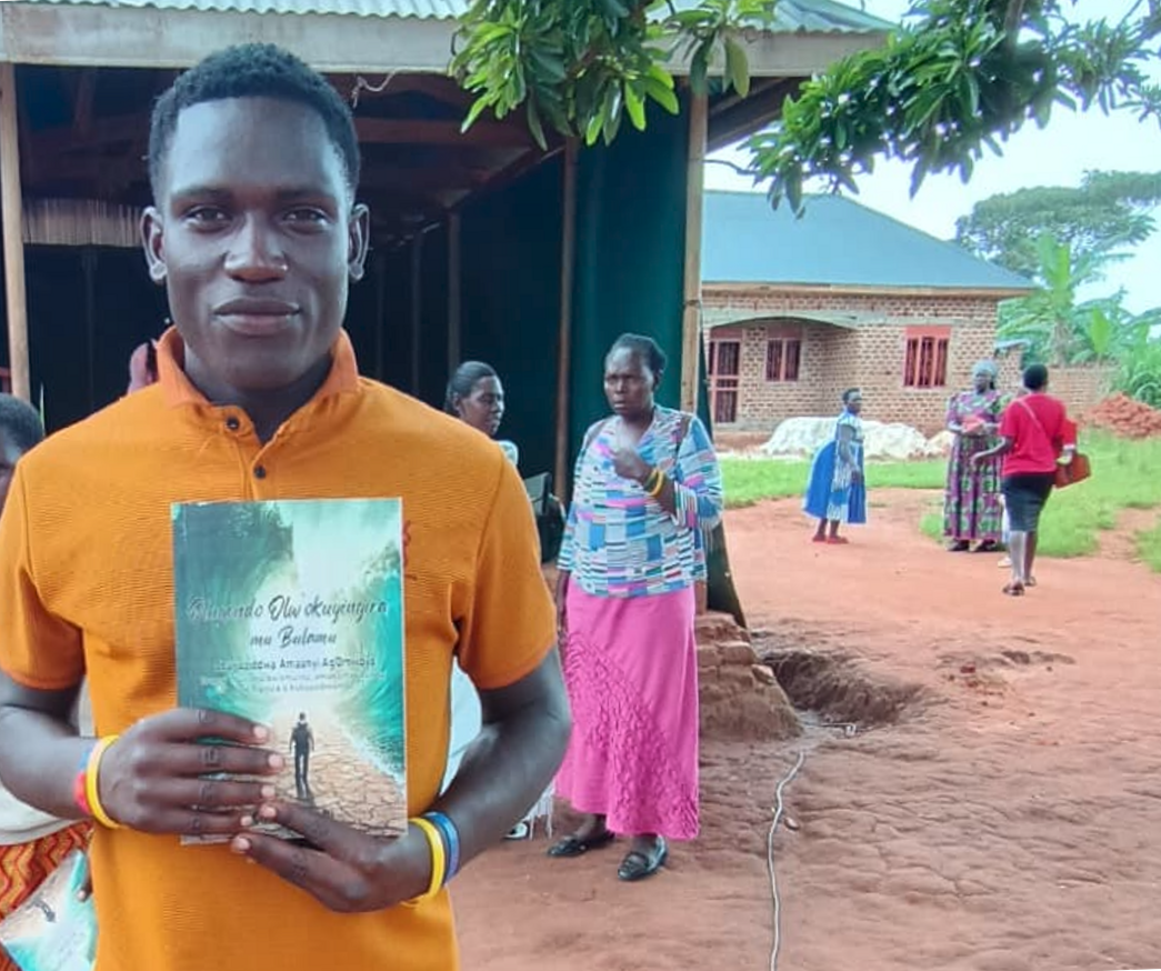 A young man in an orange polo shirt holds up a translated edition of Journey into the Spirit-Empowered Life outside a church building, with other people standing on the red earth path behind him.