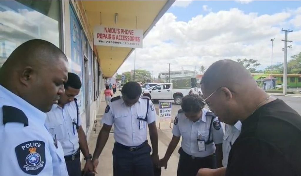 Police officers in Fiji join hands with a church leader and bow their heads in prayer on a street.