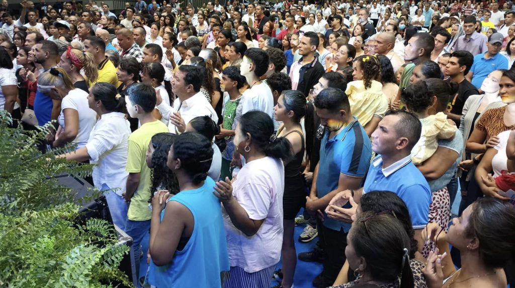 Hundreds of people stand shoulder to shoulder in prayer at a 40 Days of Love gathering in Colombia, many with hands clasped or lifted.