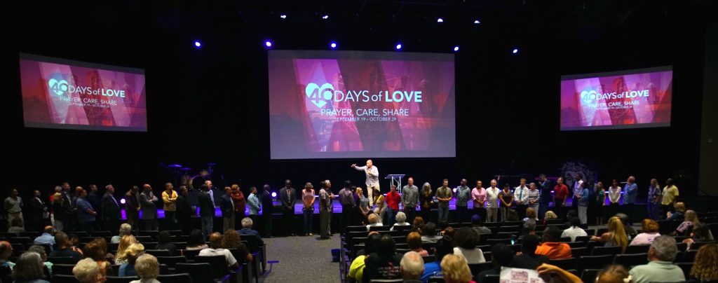 Leaders line the front of an auditorium at a 40 Days of Love launch event, with the 40 Days of Love Prayer, Care, Share branding on three large screens.