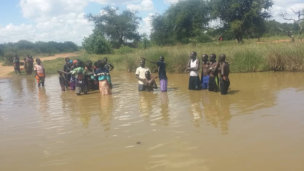 New believers wade into a river with a pastor for baptism while others watch from the bank.