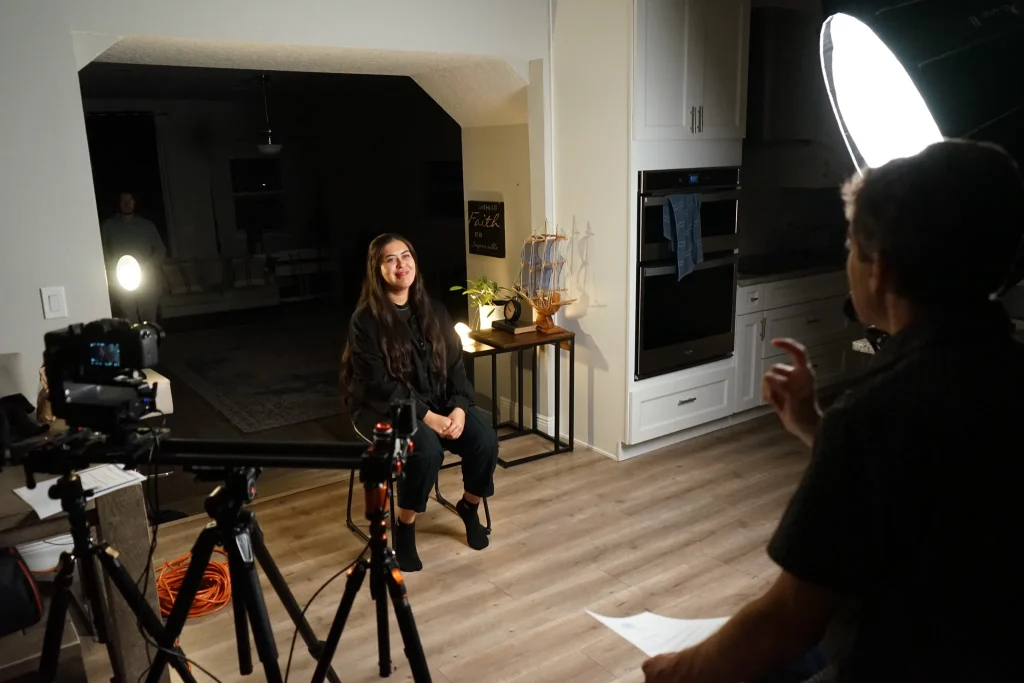 Behind the scenes on a Messiah's Story shoot: a woman shares her testimony between camera rigs while a crew member directs beside a softbox light.