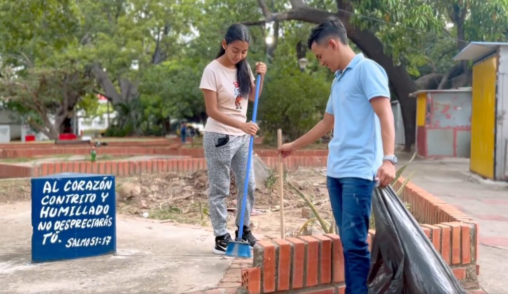 Two volunteers sweep and bag debris in a Valledupar park beside a hand-painted blue block carrying a verse from Psalm 51.